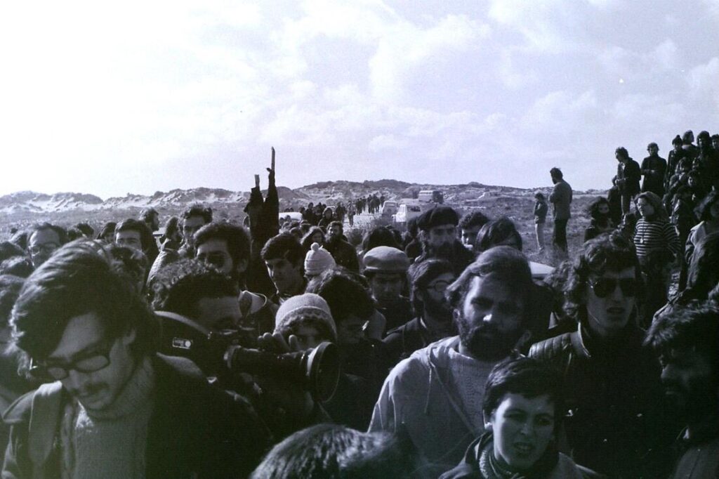 A crowd of people has gathered in the foreground. They were taking part in an anti-nuclear demonstration in Portugal in 1978. Among them, a man with a movie camera can be seen. The dune landscape of Ferrel can be seen in the background.