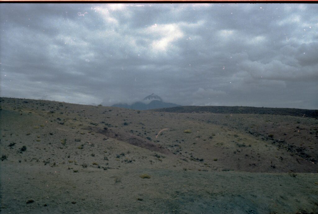 Scanned paper copy of a color photograph showing a dry, cloudy and vast landscape. A mining camp situated in a low valley is surrounded by barren mountains.